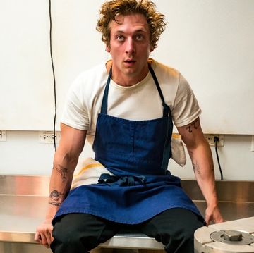 the bear, jeremy allen white sitting on top a metal work surface in a blue chef apron, looking stressed