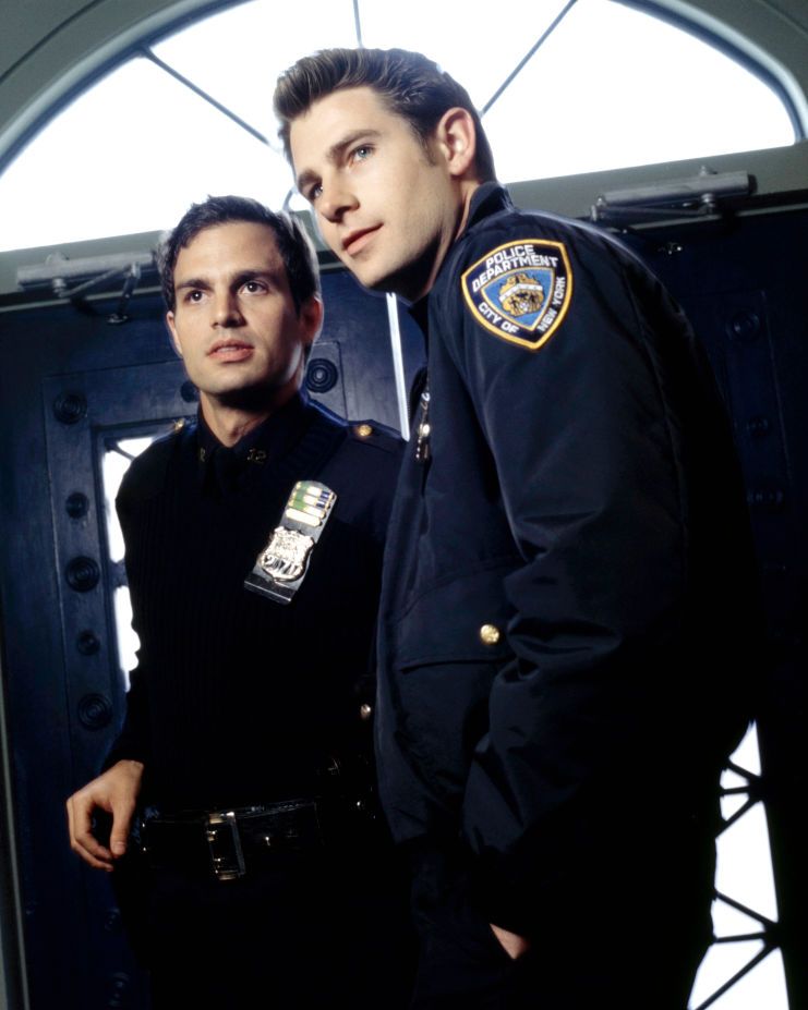 mark ruffalo and derek cecil in character for the beat, both men wear navy blue police uniforms with badges on their chests, they stand inside a set of double doors and look past the camera