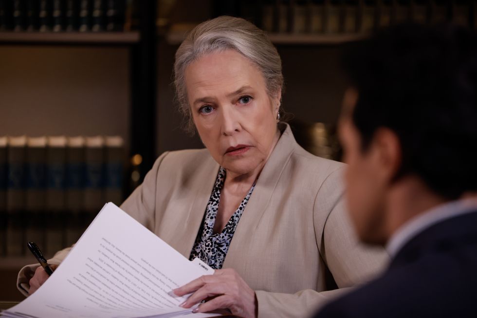 a woman in a light brown suit jacket and a black and gray patterned shirt flips through paperwork and looks at a person facing her on the right