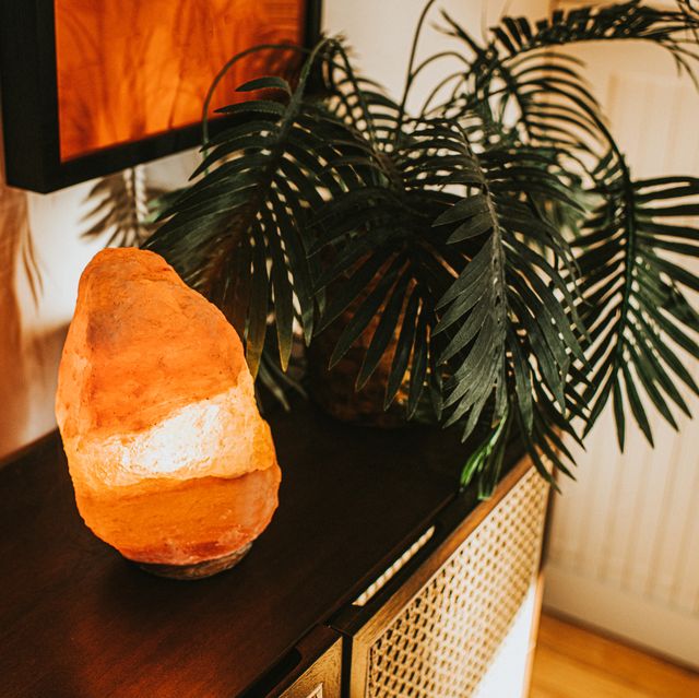 an orange himalayan salt lamp in a domestic environment beside a house plant it is on, giving a warm, soft glow