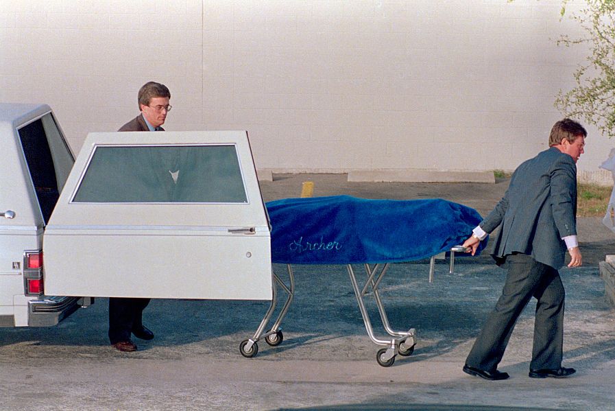 two men unloading a gurney with a covered body on top of it from a car