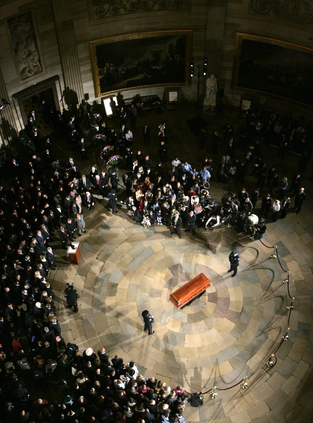 a brown casket sits in the center of a stone floor as two guards stand on either end, a large group of people observes from several feet away and a rope barrier is set up to the right