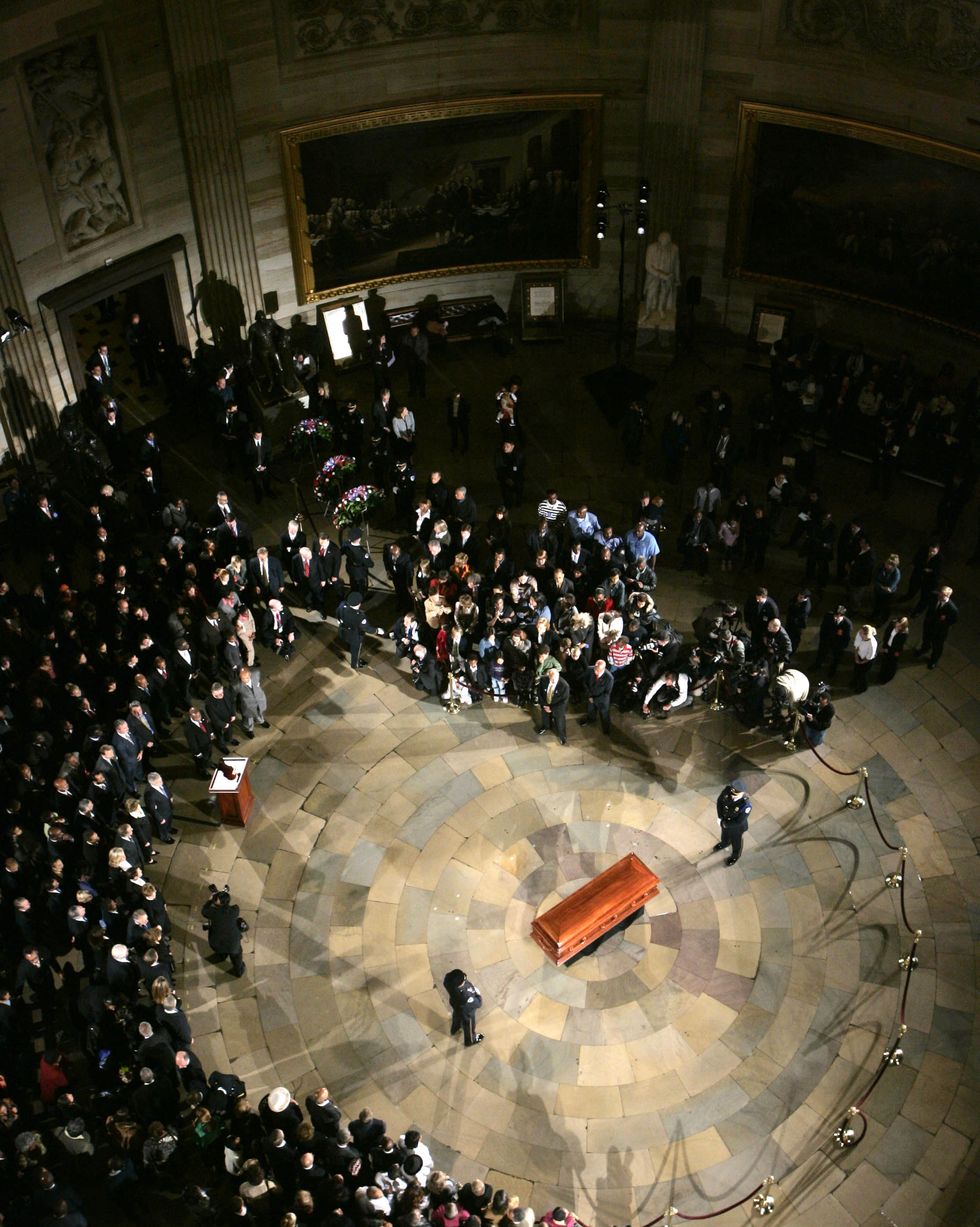 a brown casket sits in the center of a stone floor as two guards stand on either end, a large group of people observes from several feet away and a rope barrier is set up to the right
