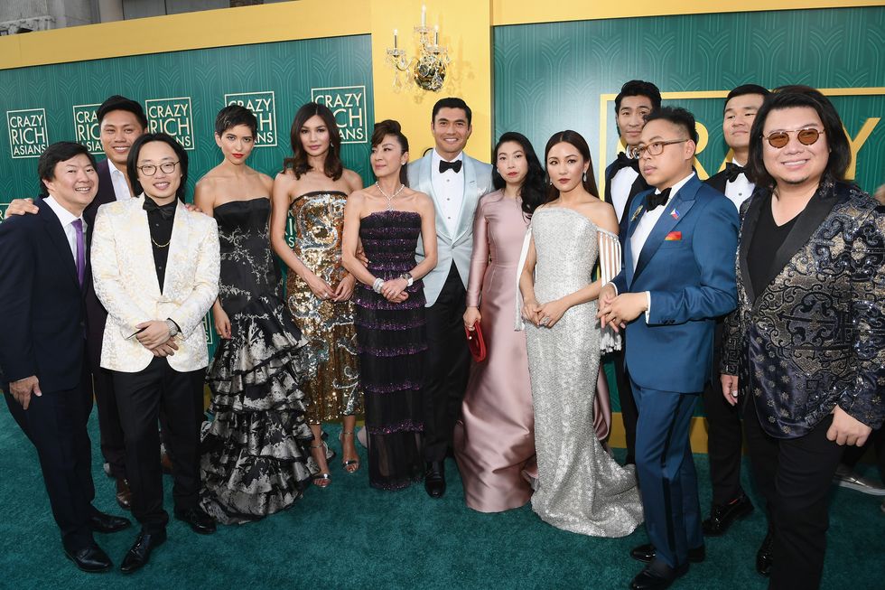 thirteen people wearing suits and dresses standing together and smiling for a photo in a ball room with green walls and several logos for the crazy rich asians on the walls