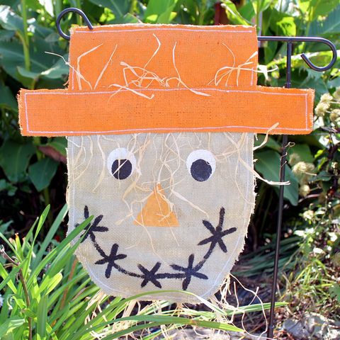 scarecrow head garden flag made out of burlap with orange hat, black and white eyes, orange triangle nose and smile made out of xs