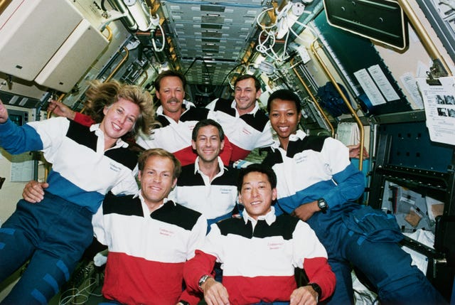 five men and two men smile for a photo while wearing matching striped polo shirts and navy pants, they float inside a space shuttle filled with equipment