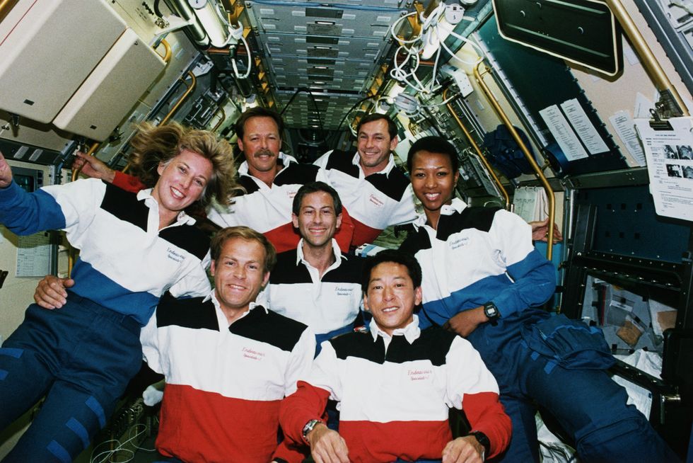 five men and two men smile for a photo while wearing matching striped polo shirts and navy pants, they float inside a space shuttle filled with equipment