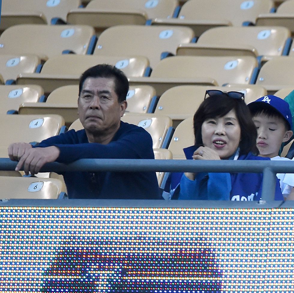 toru and kayoko ohtani sit in baseball stands, toru has his hands on a railing in front of him and wears a long sleeve navy blue shirt, kayoko wears blue and sunglasses on her head, a set of clasped hands likely belonging to an out of view child are near her face