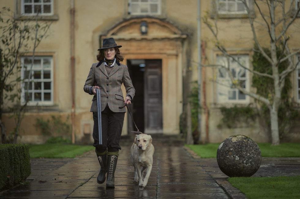 joely richardson en los caballeros, una mujer vestida con ropa de campo pasea a un perro frente a una mansión y lleva una escopeta