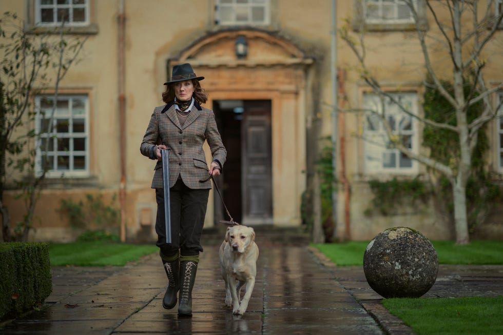 joely richardson en los caballeros, una mujer vestida con ropa de campo pasea a un perro frente a una mansión y lleva una escopeta