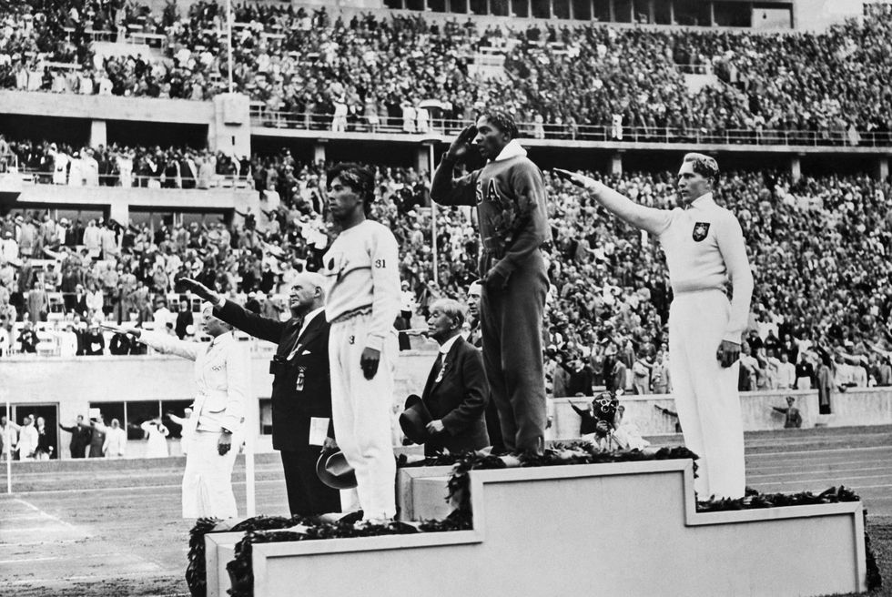 three men stand on a two tiered podium, jesse owens is in the middle and gives a salute, a crowd is in the grandstand in the background, some people give the nazi salute