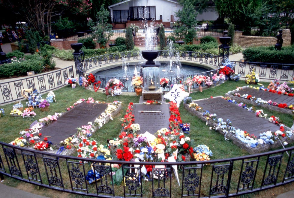 four gravestones are surrounded by flowers and gifts within a fenced in plot of grass in front of a fountain