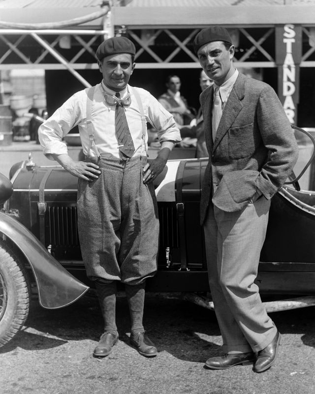 italian driver nando minoia and enzo ferrari stand in front of a car and look at the camera, minoia is in a racing outfit with goggles hanging from his neck and a hat on, ferrari wears a suite and tie with a hat