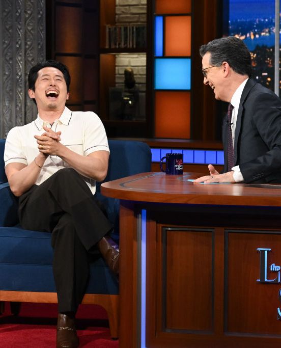 steven yeun sitting in a guest chair laughing with stephen colbert