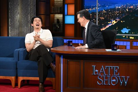 steven yeun sitting in a guest chair laughing with stephen colbert
