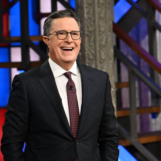 stephen colbert smiling in front of a red white and blue set piece