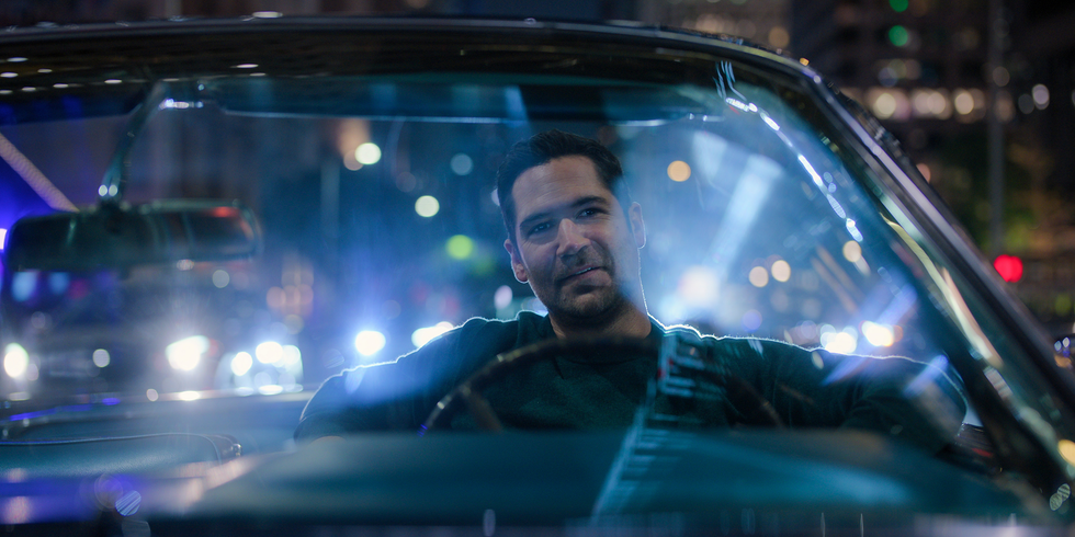 A scene inside a car at night with a person sitting in the driver's seat. The person is visible from the shoulders up and appears relaxed. The background features vibrant city lights, including blurred spotlights and street lamps, creating a lively nighttime atmosphere