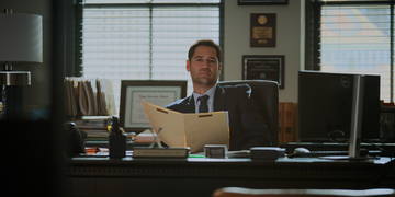 an office setting featuring a desk cluttered with various items including a computer, a stack of files, and a table lamp the individual seated in an executive chair is holding an open file folder, appearing to be engaged in thought or reviewing documents the background includes framed certificates and awards, adding a professional ambiance