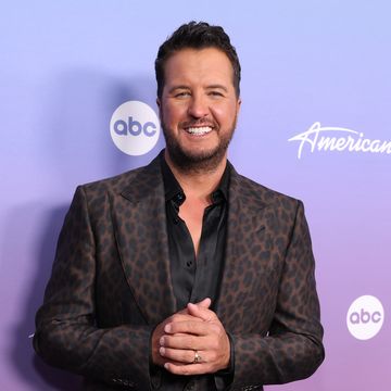 luke bryan smiles at the camera in front of a purple background, he wears a cheetah pattern suit jacket over a black collared shirt that is unbuttoned, he clasps his hands in front of his waist