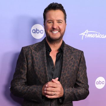 luke bryan smiles at the camera in front of a purple background, he wears a cheetah pattern suit jacket over a black collared shirt that is unbuttoned, he clasps his hands in front of his waist