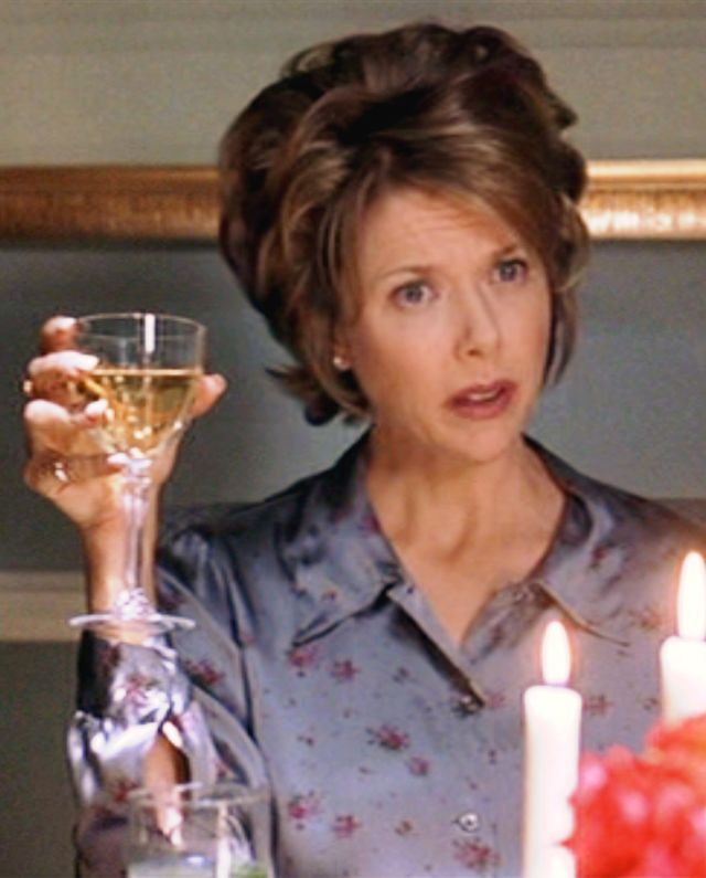 annette bening in character for american beauty, she sits at a dining table and holds a glass of wine up, in front of her are lit candles and red flowers