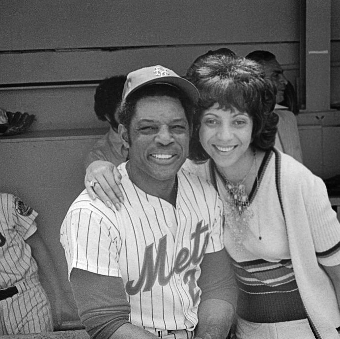 willie mays and mae mays smile at the camera from a baseball dugout, willie wears a new york mets jersey and baseball cap, mae wears a striped sweater set