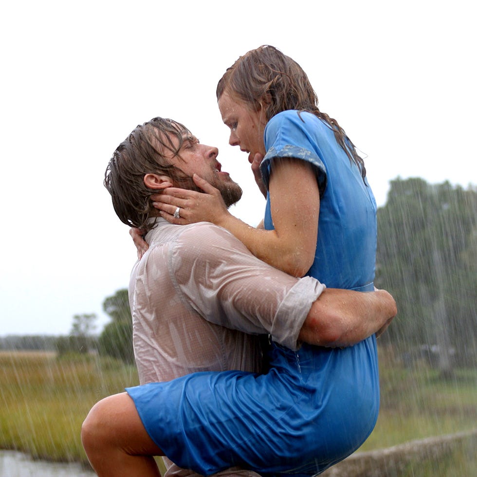 a couple is caught in a romantic moment amidst heavy rainfall, the woman is wearing a blue dress and is lifted by the man, who is wearing a wet shirt