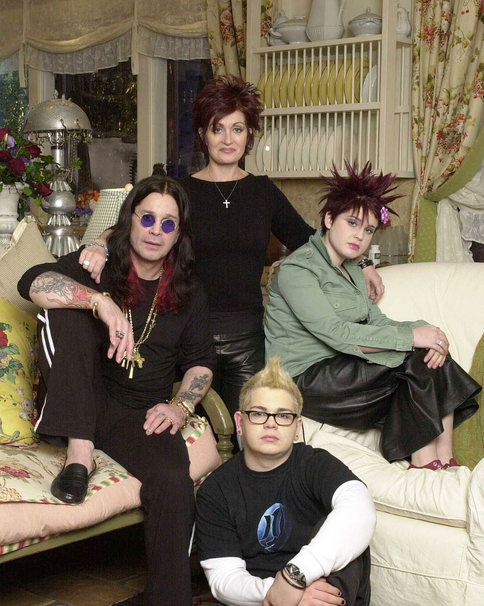 a woman stands behind a man and two children who are seated on furniture and the floor, all four people look at the camera