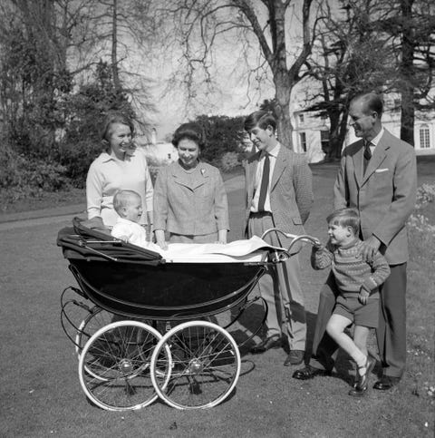princess anne, queen elizabeth ii, prince charles, prince andrew, and prince philip stand around a stroller that prince edward sits inside, everyone is smiling