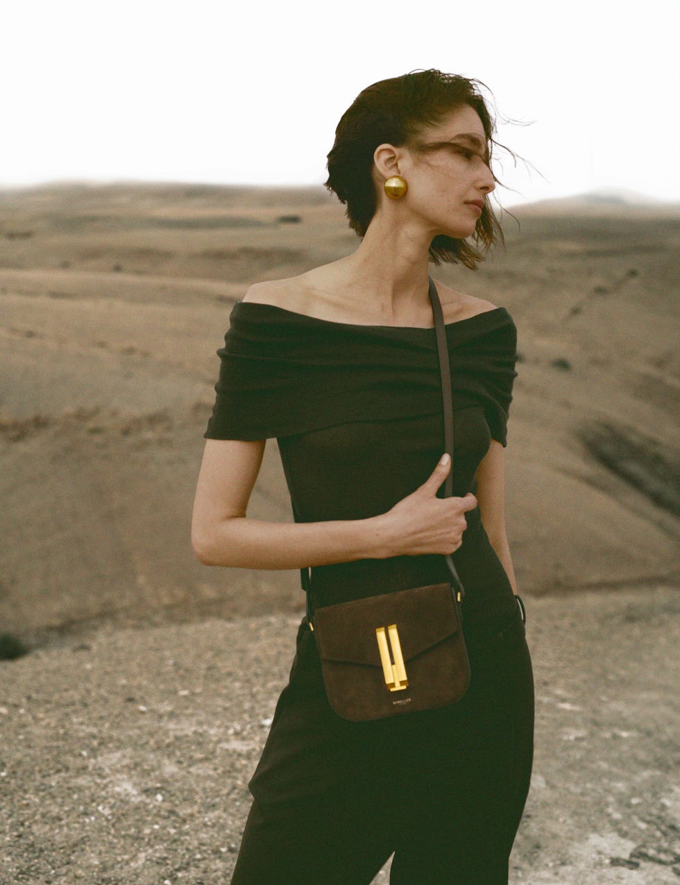 a stylish person wearing a black outfit stands with a handbag in a desert landscape