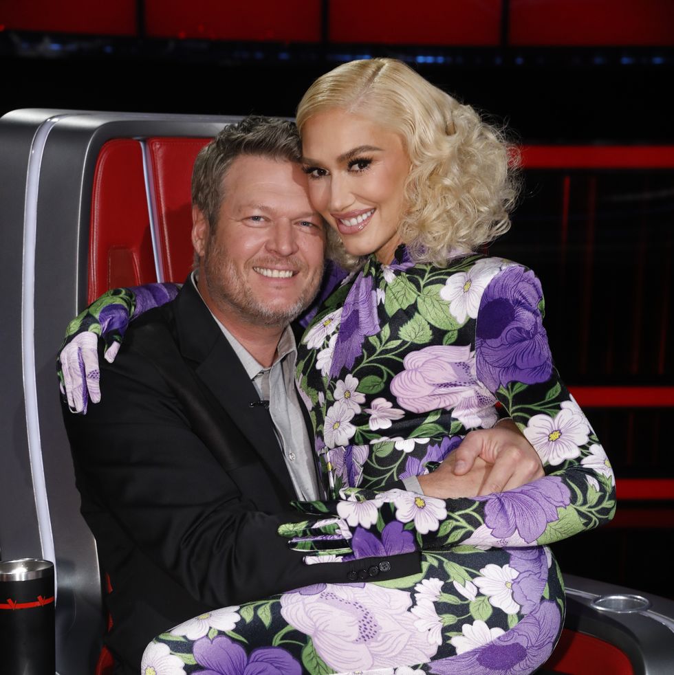 blake shelton sits in a red coach chair from the voice, gwen stefani sits on his lap, they hug each other and smile at the camera