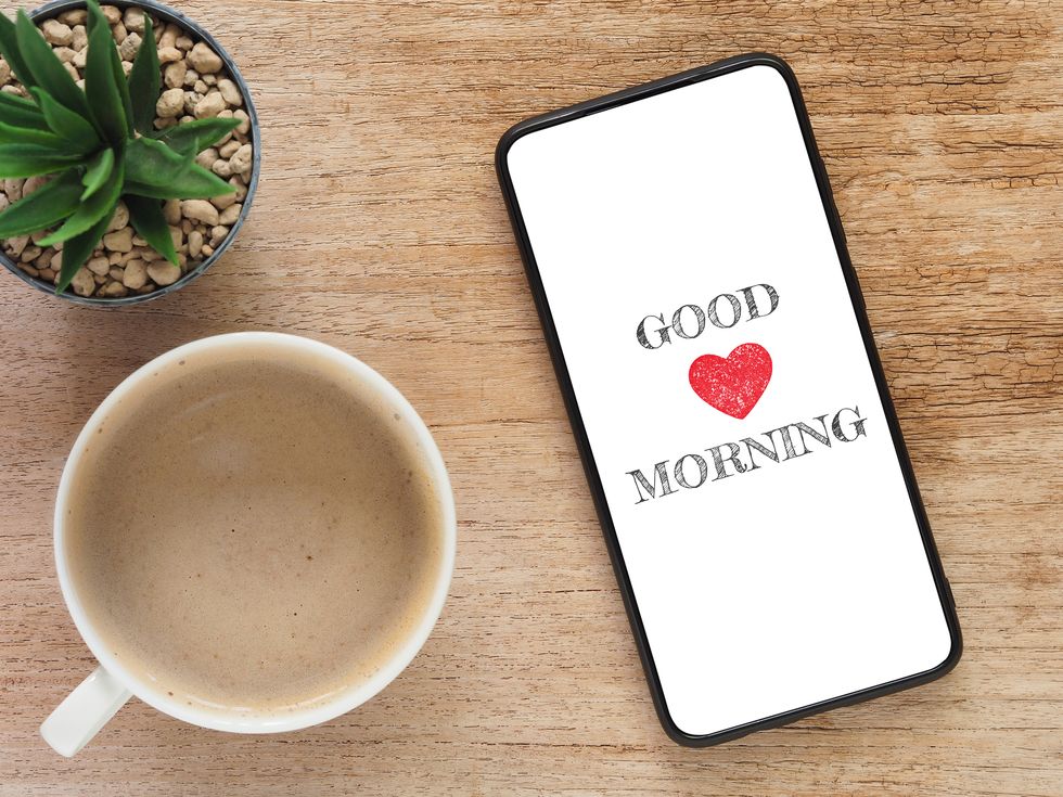 the word "good morning" with a red heart on the screen smartphone and coffee in a white mug, cactus on a wooden background top view, flat lay