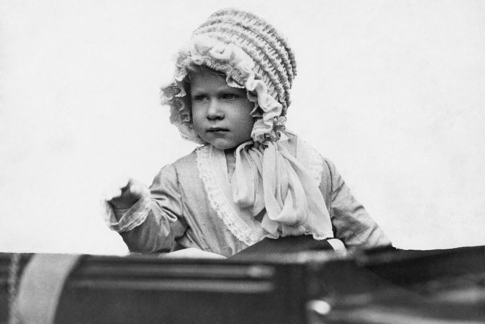princess elizabeth as a baby sits and waves, she wears a ruffled bonnet and a long sleeve dress
