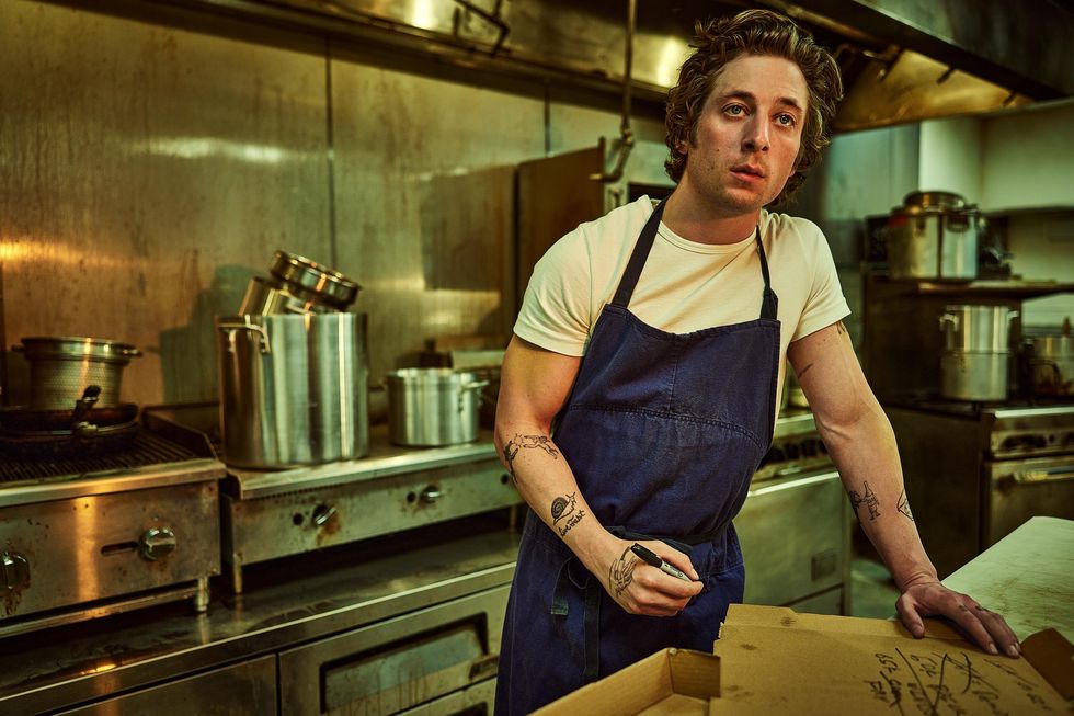 jeremy allen white stands in a commercial kitchen wearing a white tshirt and blue apron, he holds a pen in one hand and leans on a table that has a piece of cardboard resting on it
