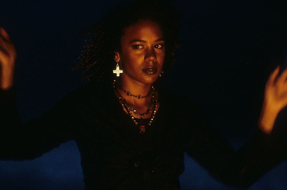 in a scene from the craft, rochelle   a young black woman   wears a black top, layered necklaces, and cross earrings as she holds her hands up to cast a spell