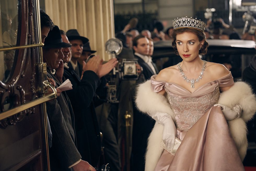 vanessa kirby in character as princess margaret for the crown, she wears a pink ballgown, white fur stole, large diamond necklace, and diamond tiara, she looks to the side and walks toward an open door, several people are watching around her
