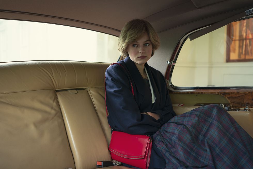 emma corrin, playing princess diana, sits inside the backseat of a car, she is wearing a long sleeve navy jacket and plaid long skirt with a pink small purse