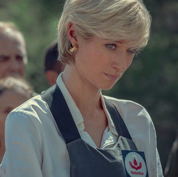 elizabeth debicki in character as princess diana for the crown, she wears a light colored collared shirt and a navy blue apron over top with gold hoop earrings, she looks past the camera with her head tilted down and a sad look on her face