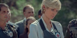 elizabeth debicki in character as princess diana for the crown, she wears a light colored collared shirt and a navy blue apron over top with gold hoop earrings, she looks past the camera with her head tilted down and a sad look on her face