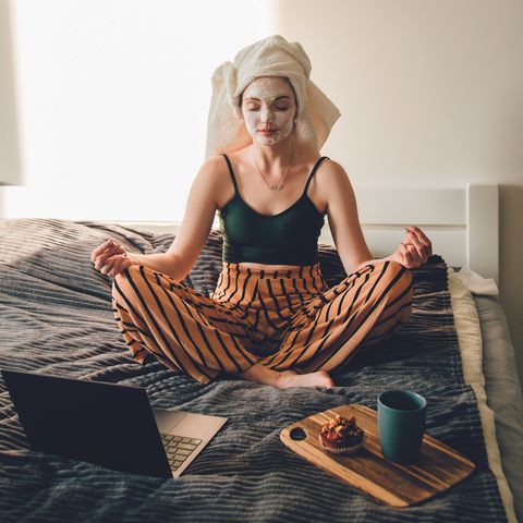 things to do by yourself woman with a face mask on meditating on her bed with a laptop and wooden board with a muffin and mug