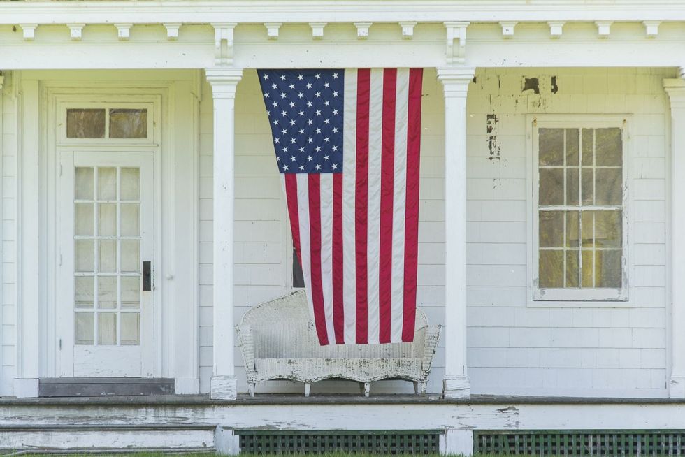 american flag hanging on the front porch of a white shingled country home for the 4th of july