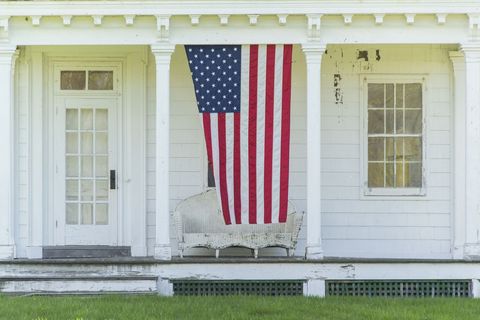 american flag hanging on the front porch of a white shingled country home for the 4th of july