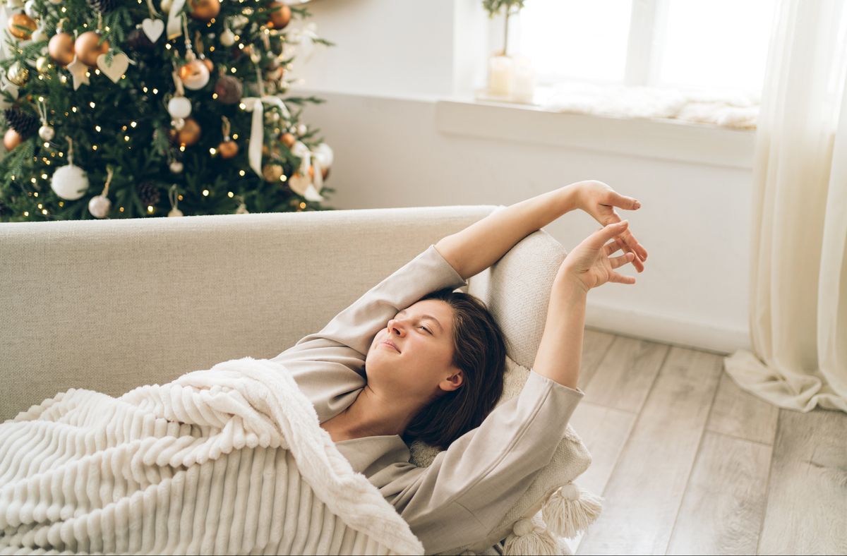 young beautiful woman is stretching lying on sofa under blanket in light interior with decorated christmas tree and many lights concept of new year holidays photography from above