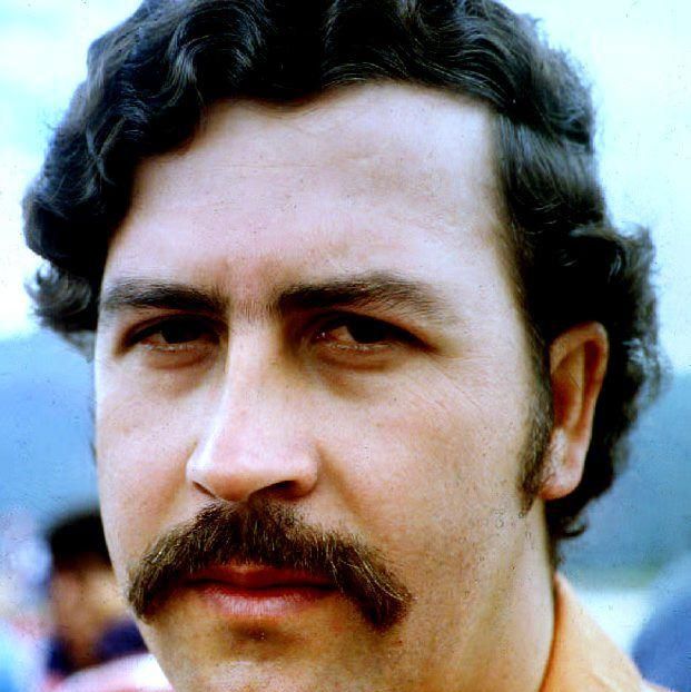 pablo escobar looks at the camera with a neutral expression, he is wearing an orange shirt and has a bushy large mustache