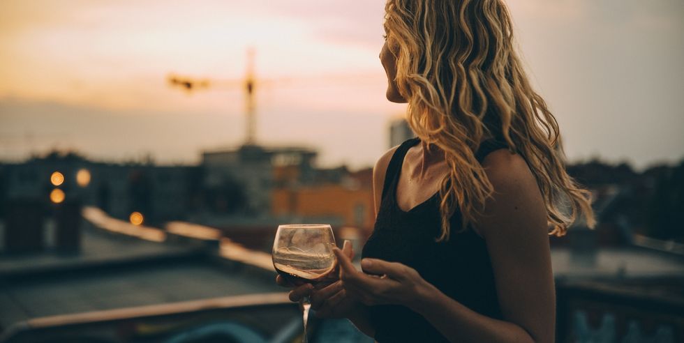 thoughtful young woman having wine while looking away on terrace during rooftop party at sunset
