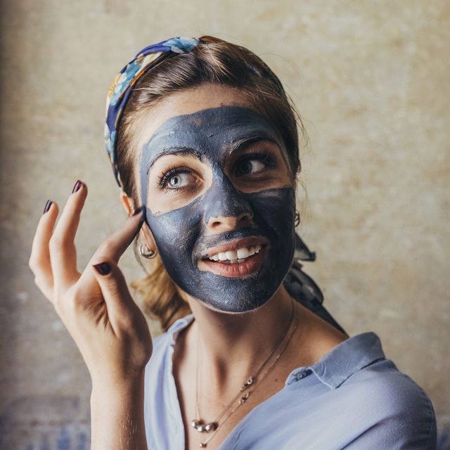 Thoughtful young woman smiling while applying facial mask against wall at home