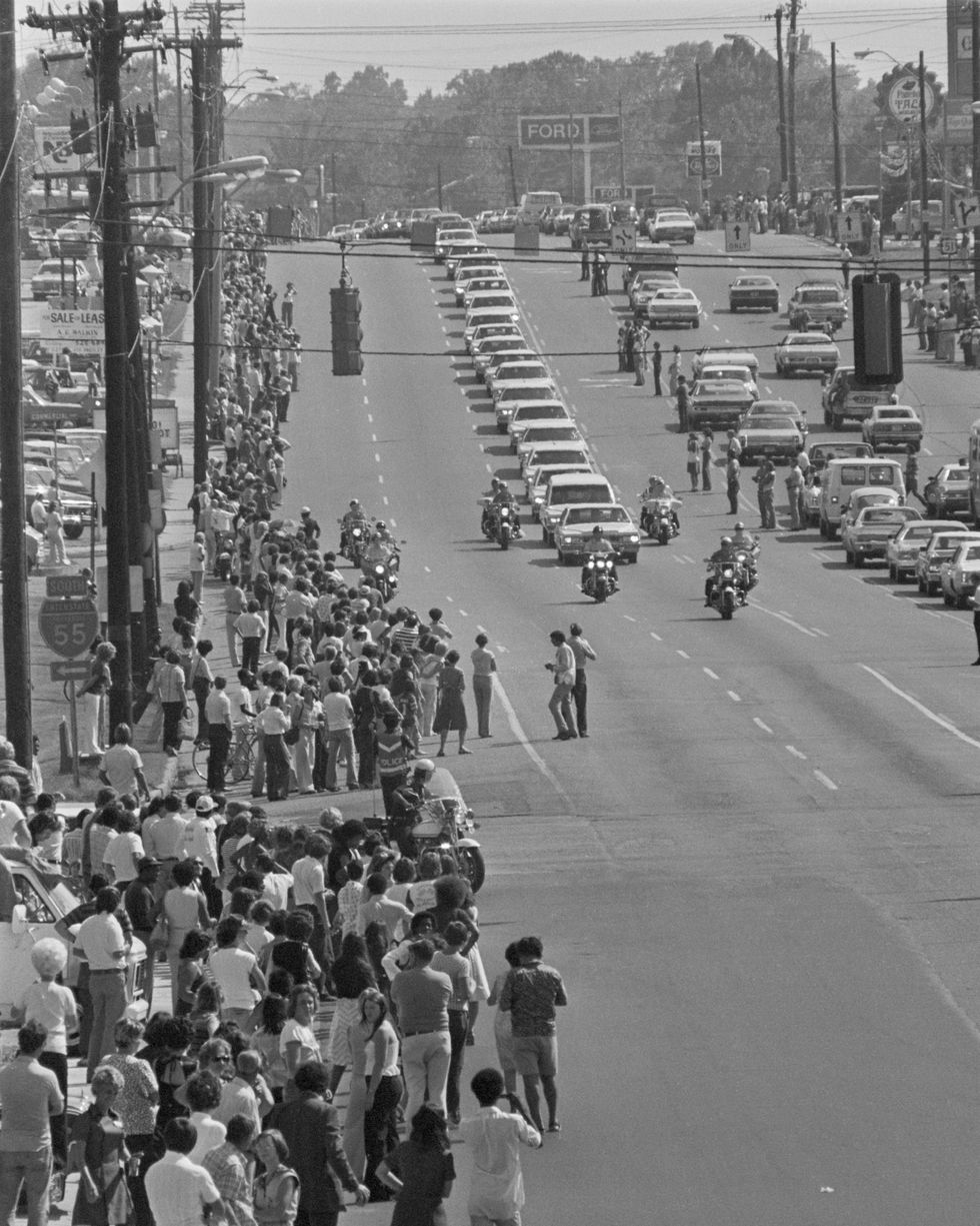 people stand along sidewalks and watch a motorcade of white cadillacs and a police escort drive by