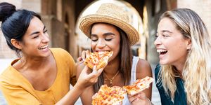 three cheerful multiracial women eating pizza in the street   happy millennial friends enjoying the weekend together while sightseeing an italian city   young people lifestyle concept