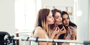 Three female teenager friends putting on lipstick in front of the mirror at home.