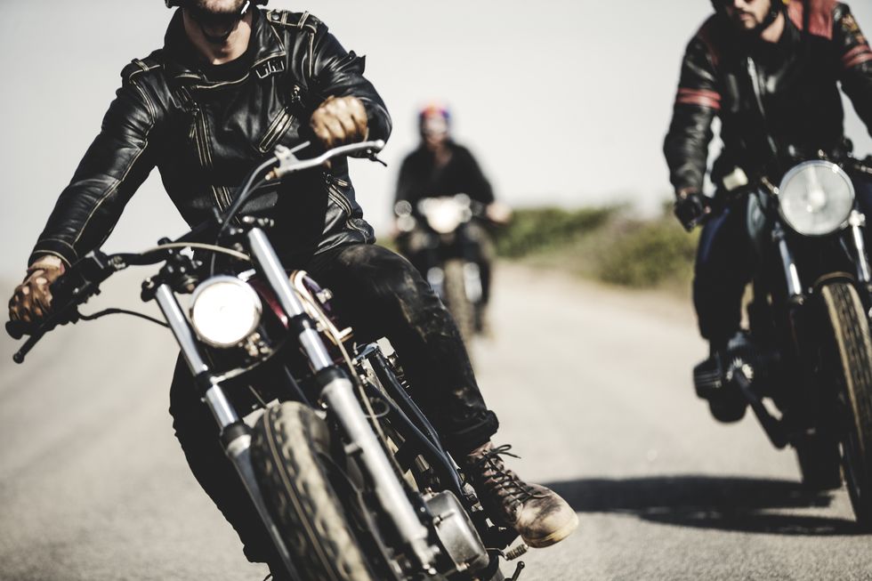 three men wearing leather jackets riding cafe racer motorcycles along rural road